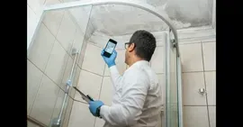A person in a white shirt and blue gloves holds a phone up to photograph water damage on a bathroom ceiling.