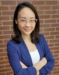 Woman with glasses in a blue blazer, arms crossed, smiling. In front of a brick wall.