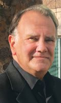 Man in black suit and shirt, smiling slightly, outdoors near a stone wall.