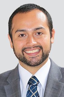 Man wearing a suit and tie smiling, with a beard, against a gray background.