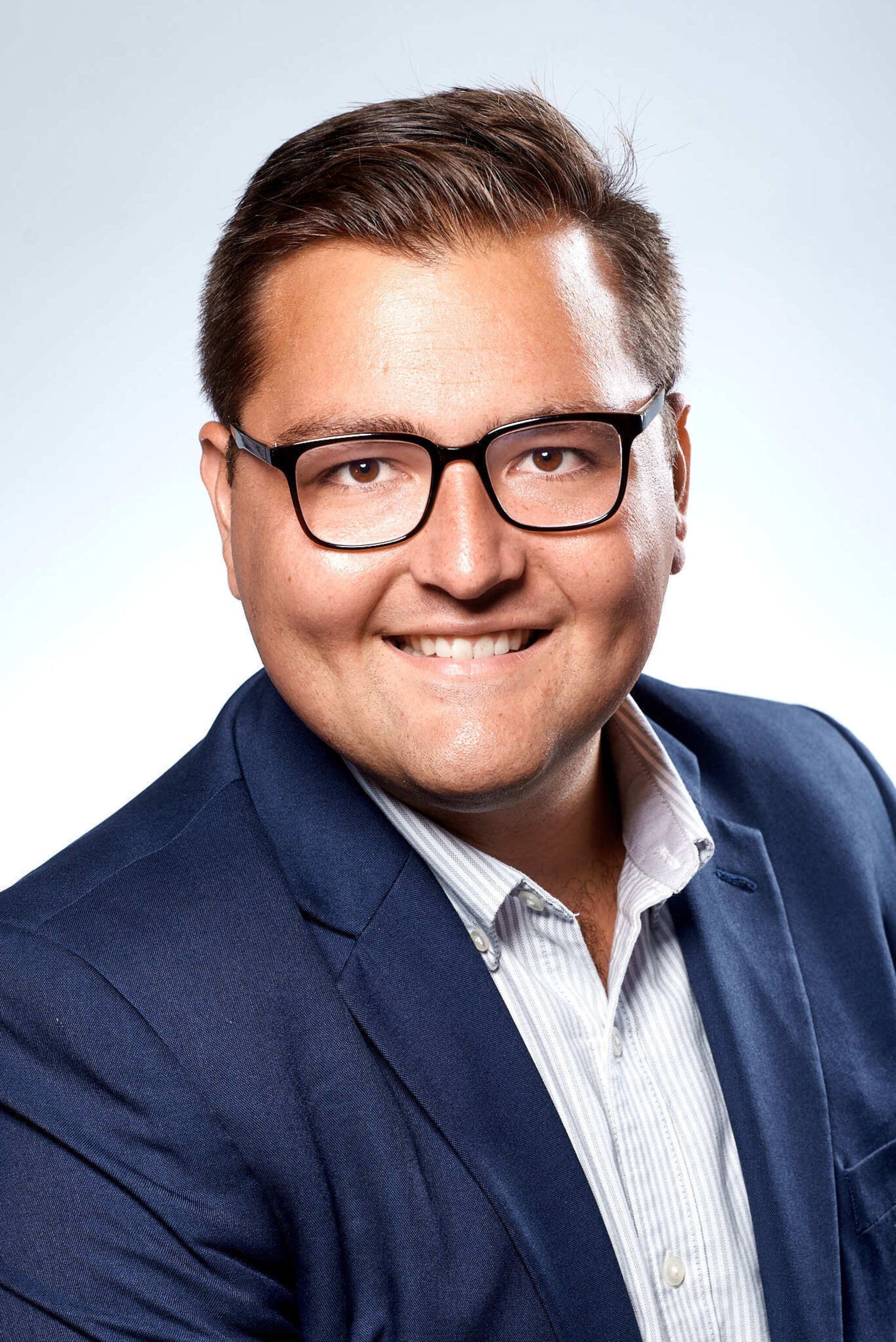 Man in glasses smiles, wearing blue blazer and light patterned shirt against a light gray background.