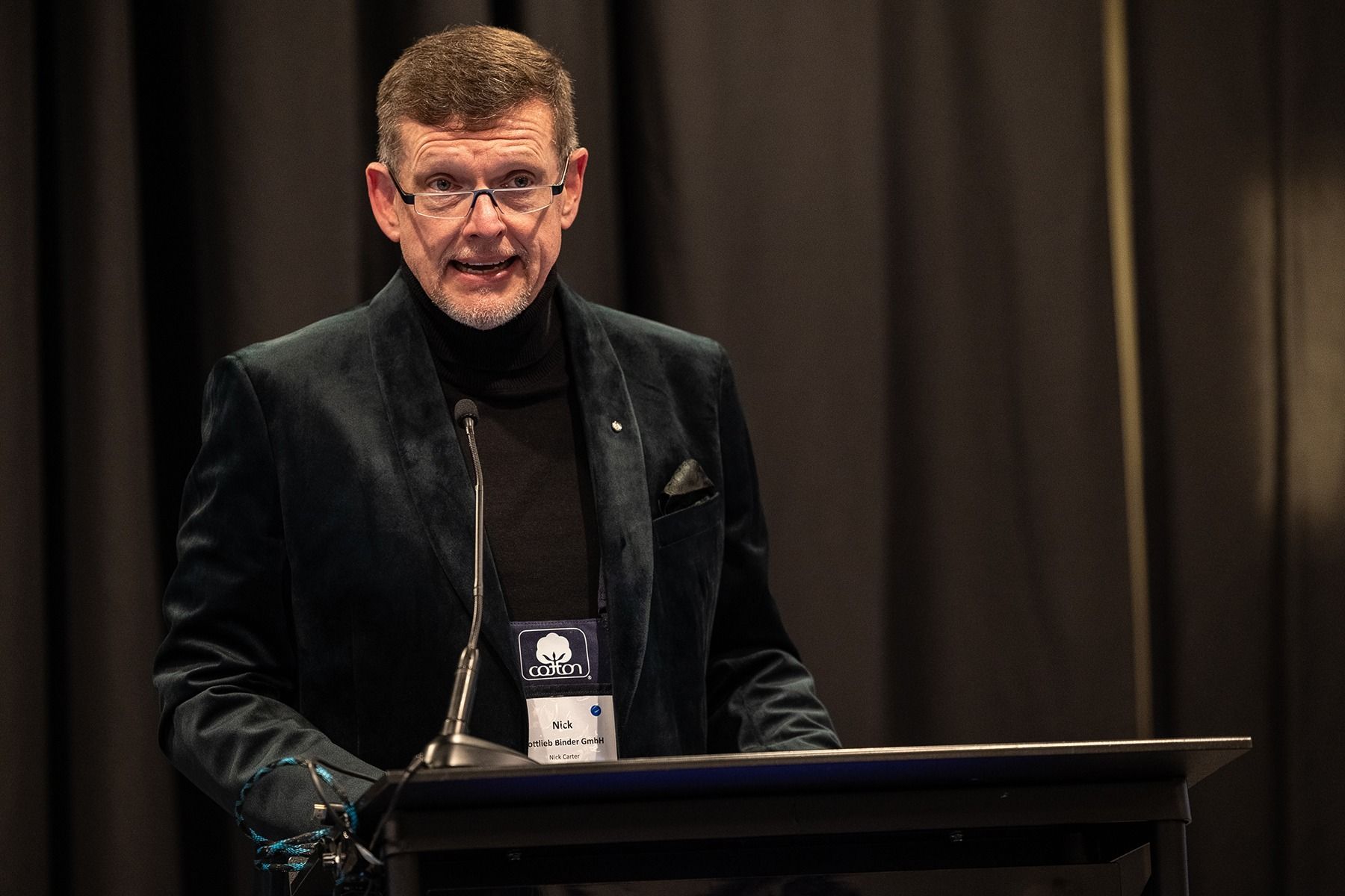 Man in dark blazer and turtleneck speaks at a podium with microphone. Dark curtains in background.
