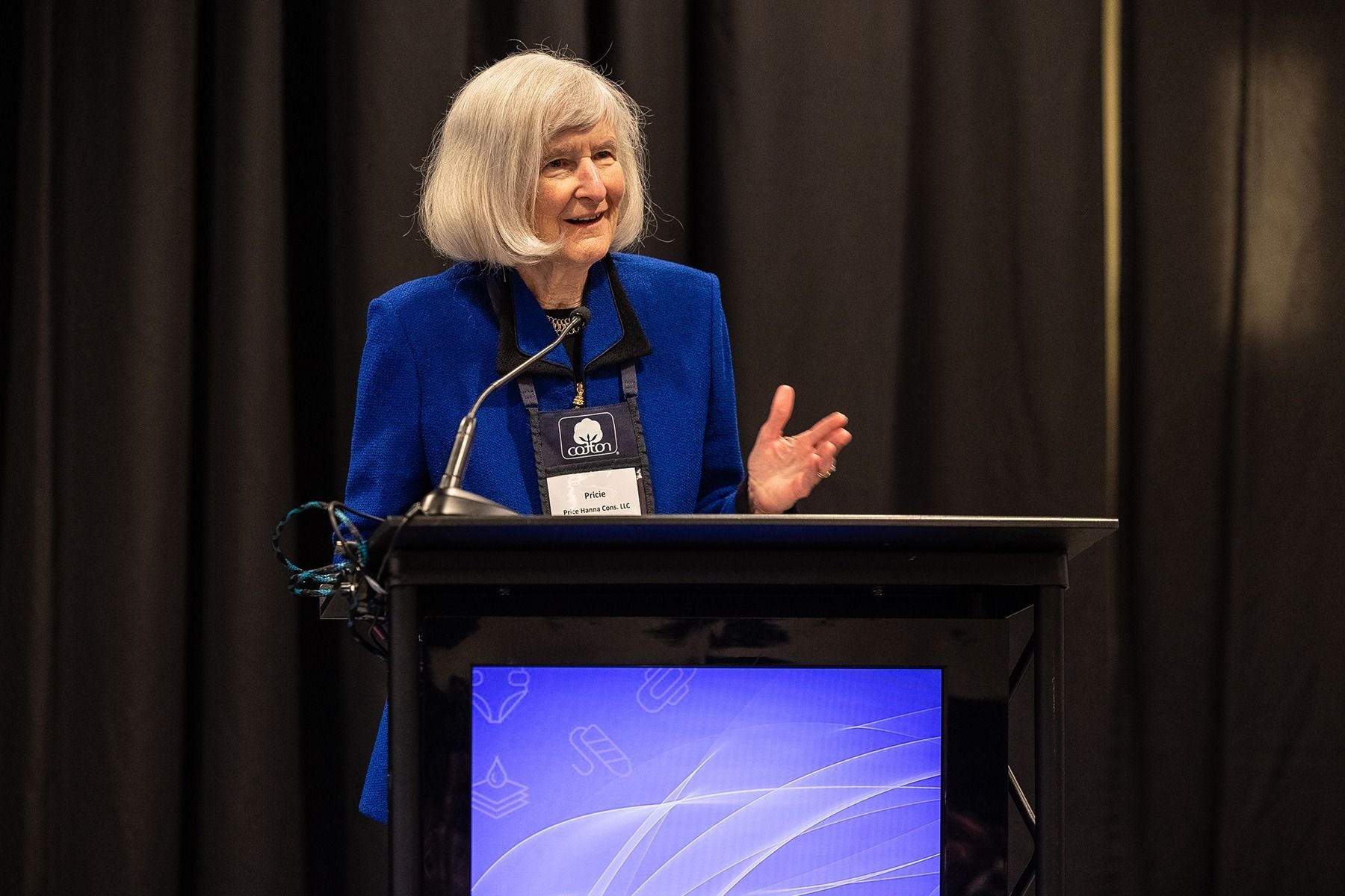 Woman speaking at a podium, wearing a blue blazer, with hands gesturing, on a stage with a screen.