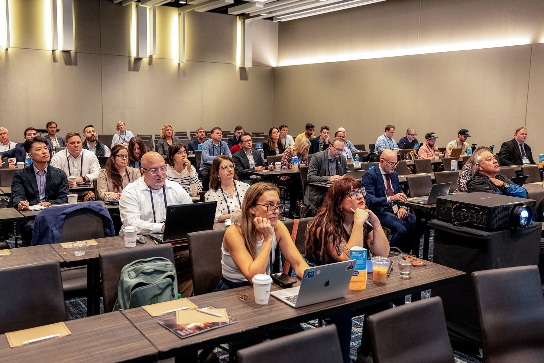 Audience in conference room, some with laptops, listening to a presentation. Tables, projector, neutral colors.