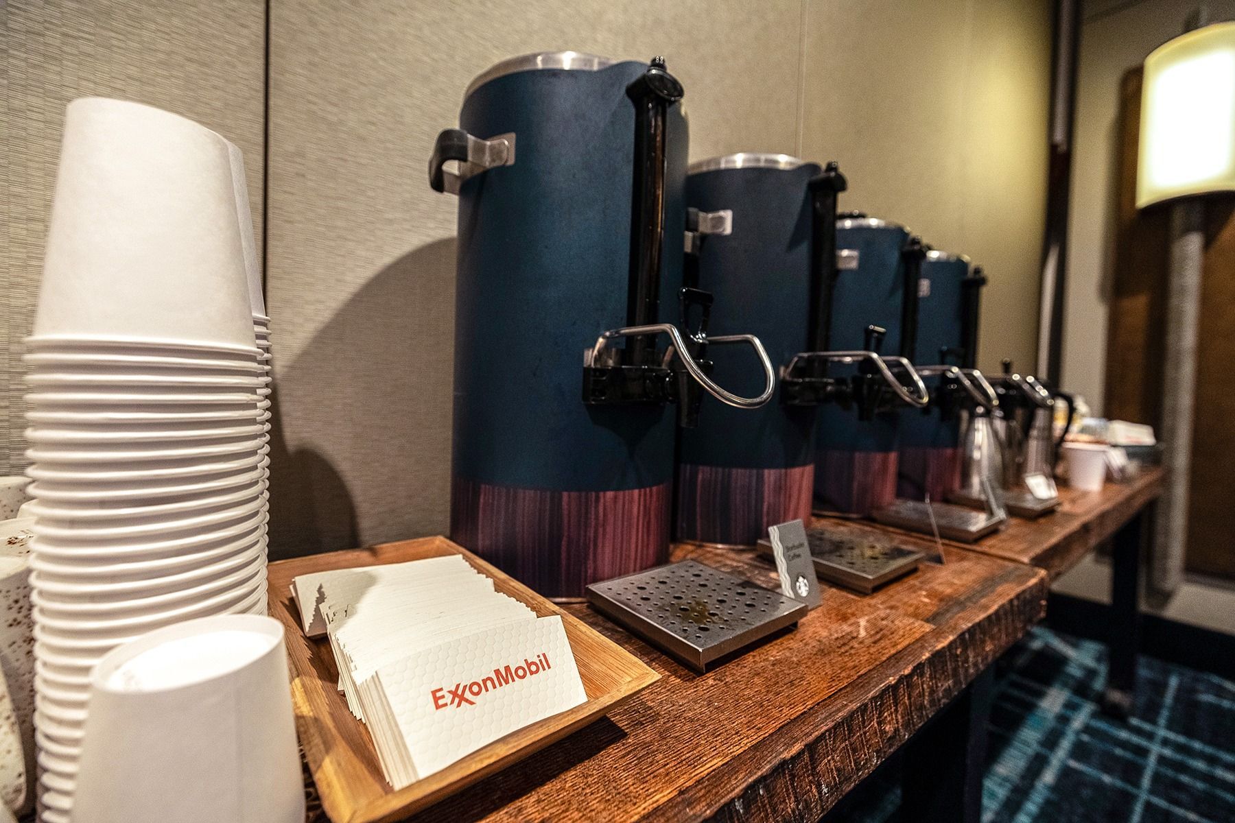 Coffee station with stacked white cups, coffee urns, and napkins on a wooden table.