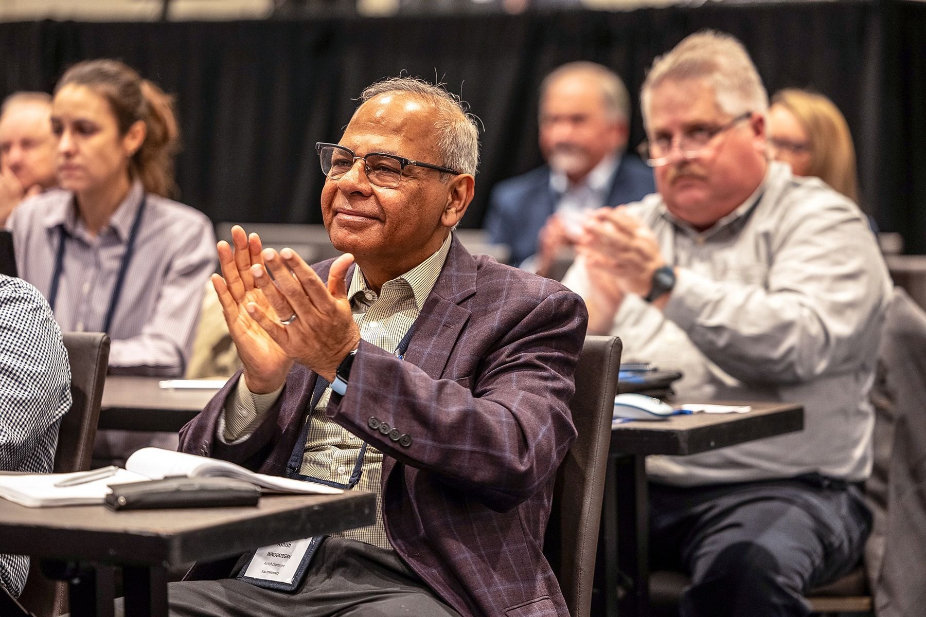 Man in blazer applauding at a conference table, others visible in background.