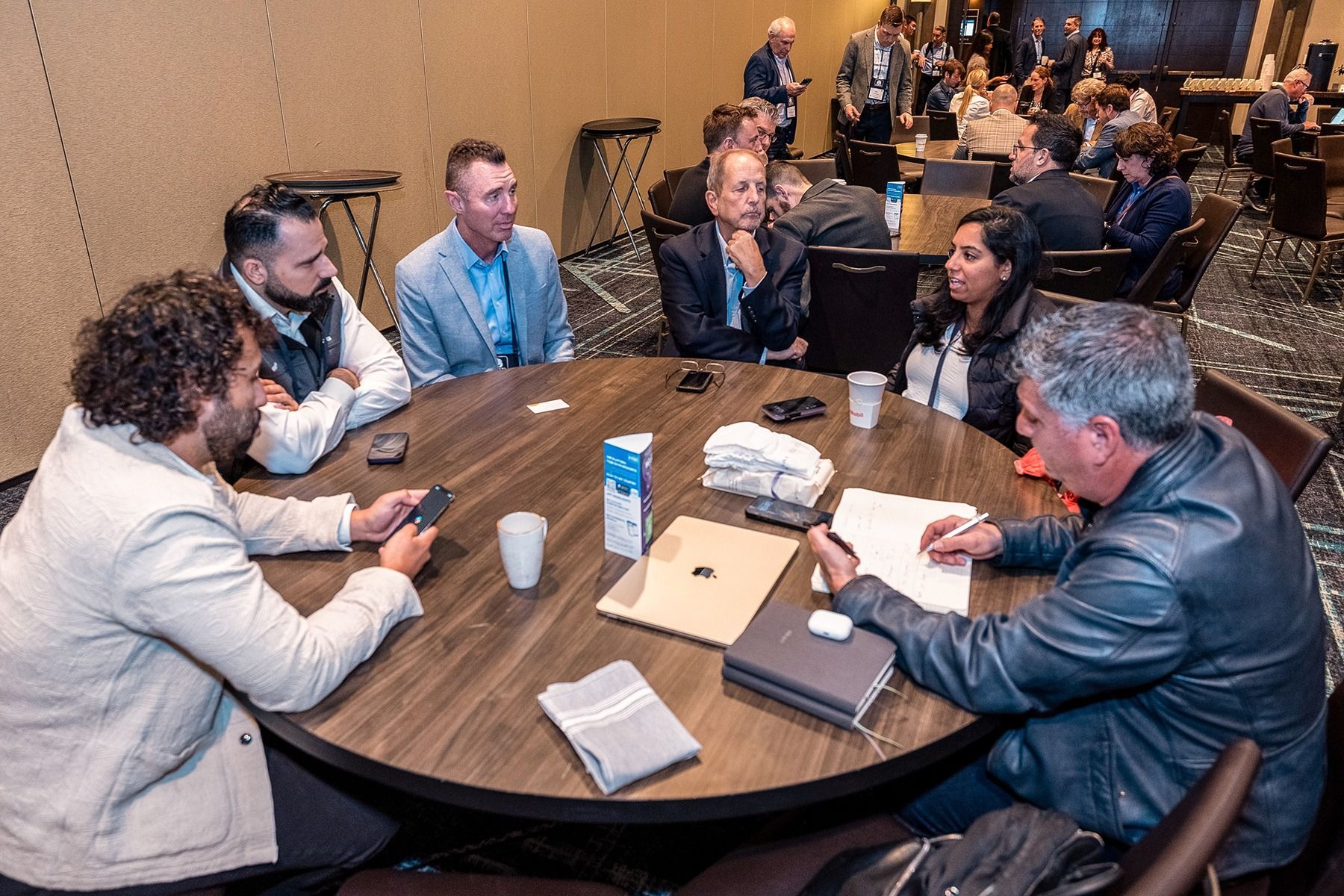 People seated at a round table, possibly in a meeting. Some are looking at papers, others at phones.