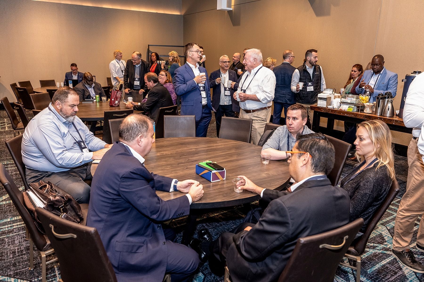 People gathered at round tables in a conference room, socializing. Some are seated, while others stand and chat.