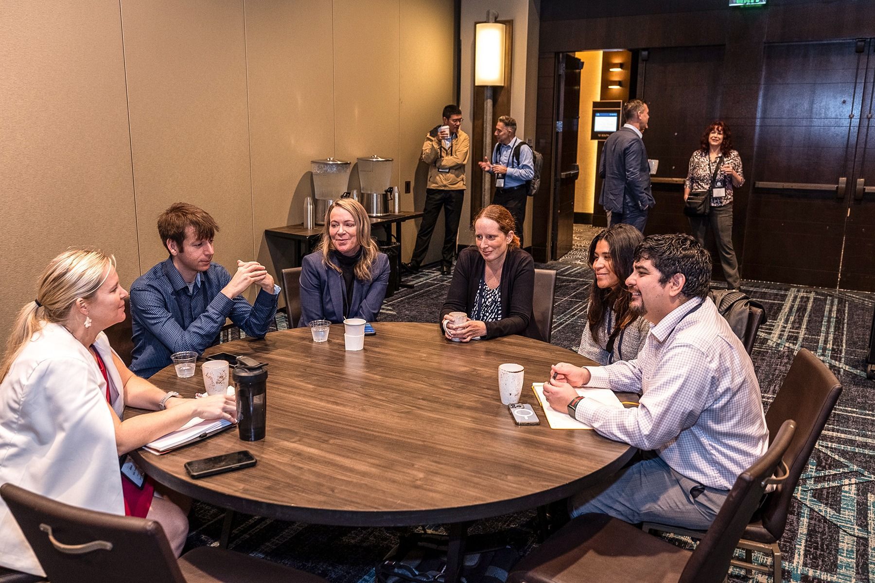 Group of people at a round table, talking. Inside a room, some in the background.