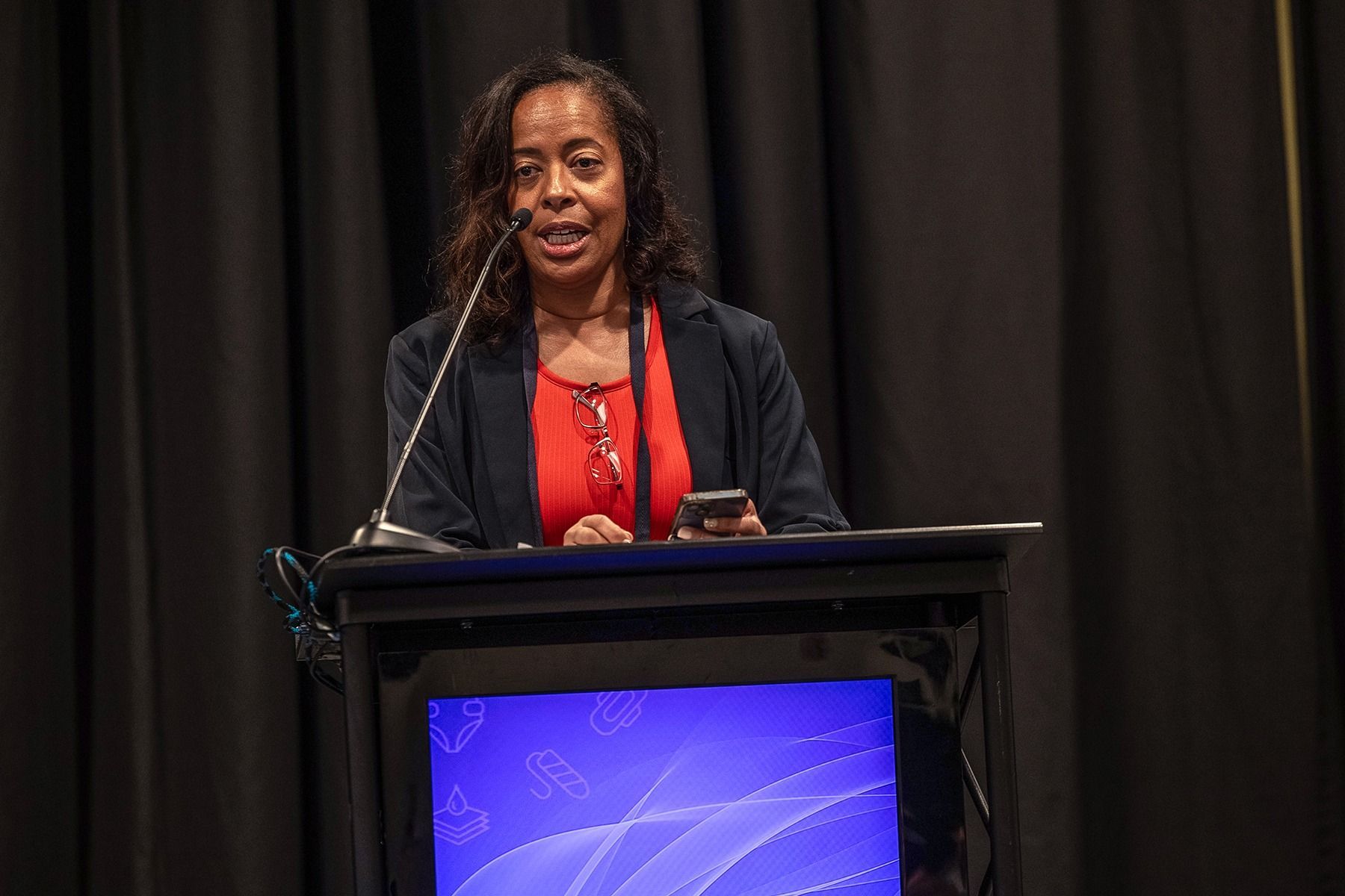 Woman in red top and blazer speaking at a podium with a screen against a dark curtain.