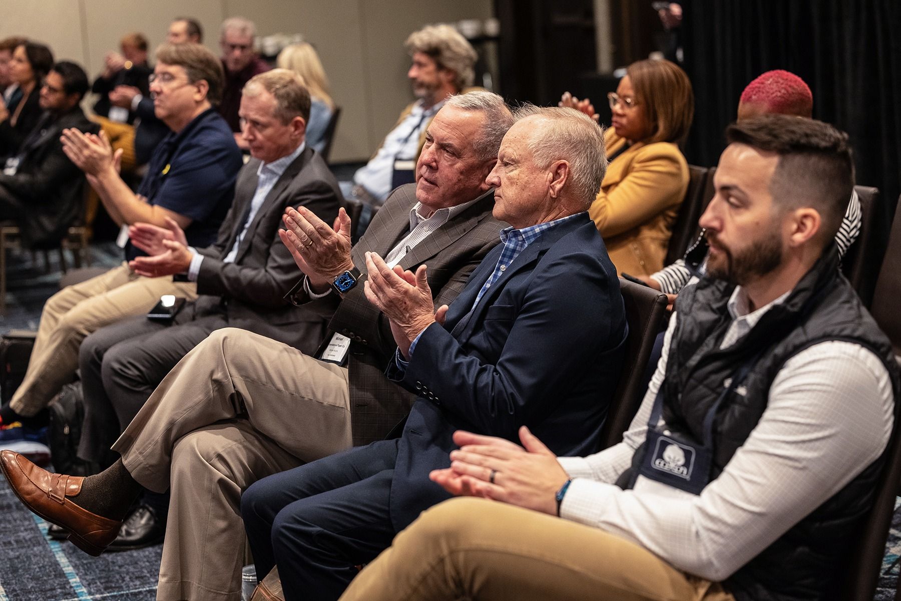 Audience applauds at a conference, men in suits and casual attire sit and clap in a hotel meeting room.