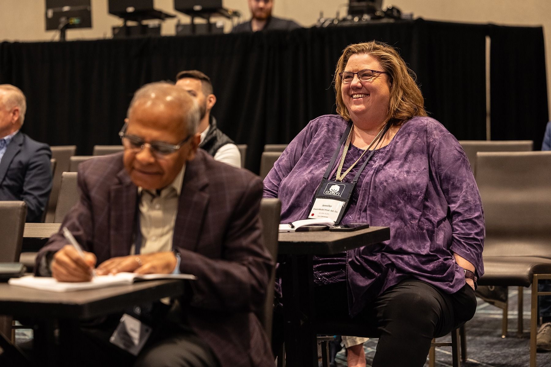 Woman in purple smiling at a conference. Man writing notes in the foreground.