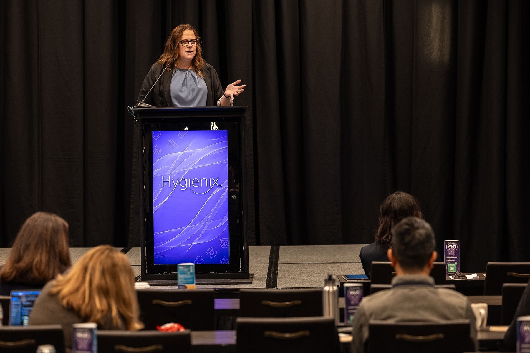 Woman presenting at a conference, standing at a podium with a screen, facing an audience.