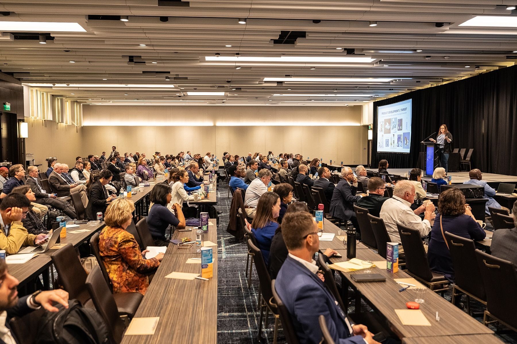 A conference room with a speaker presenting to a large audience seated at long tables.