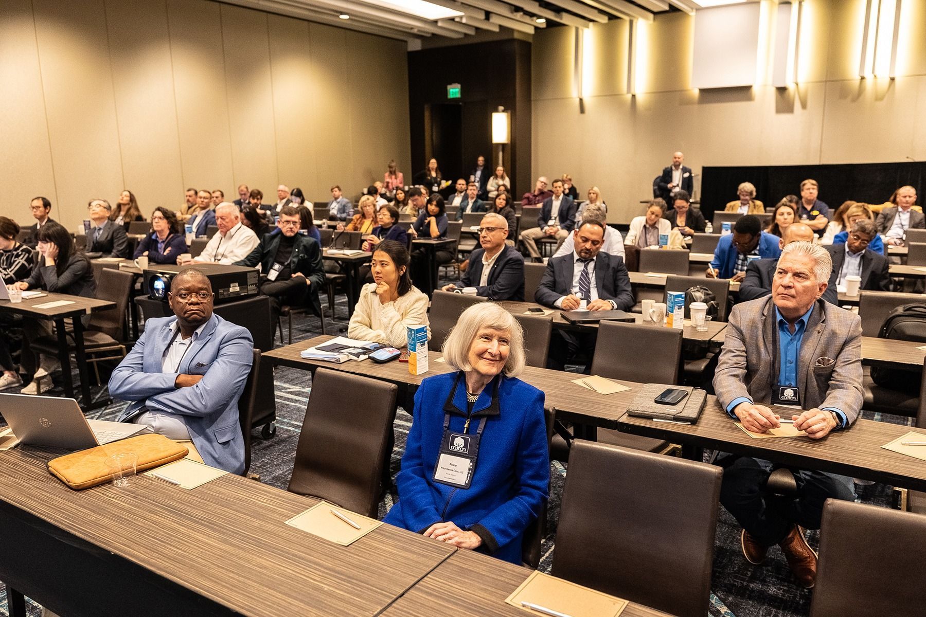 Audience in a conference room; people seated at tables, listening to a presentation.