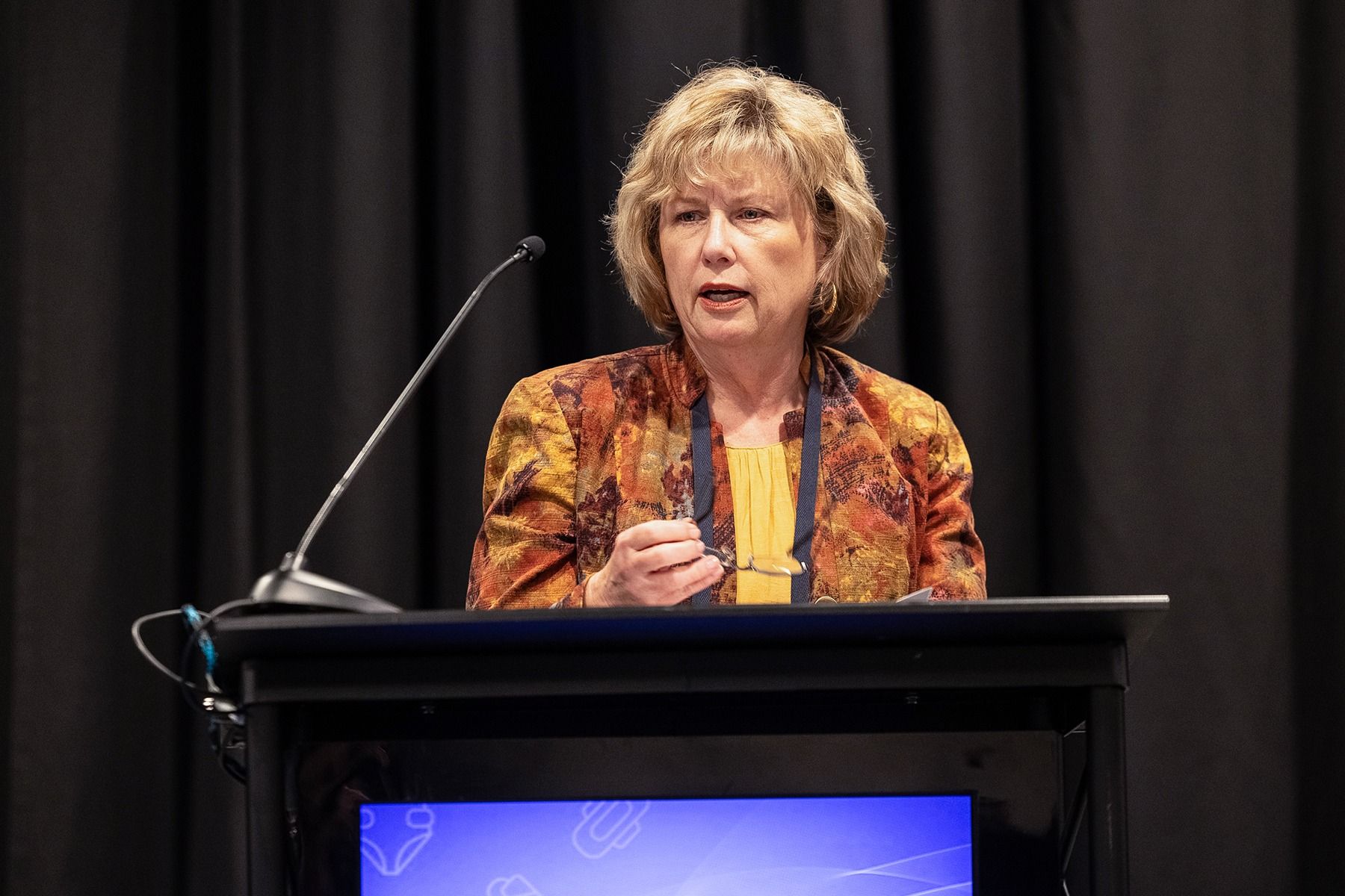 Woman speaking at a podium with a microphone, wearing a patterned orange blazer, dark background.