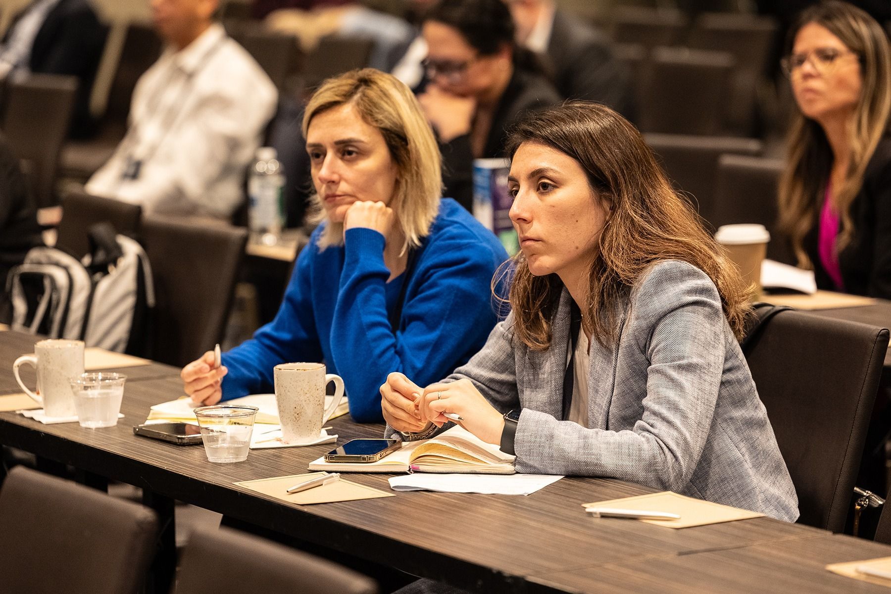 Two women seated at a conference table, listening intently. One in blue, the other in a gray jacket.