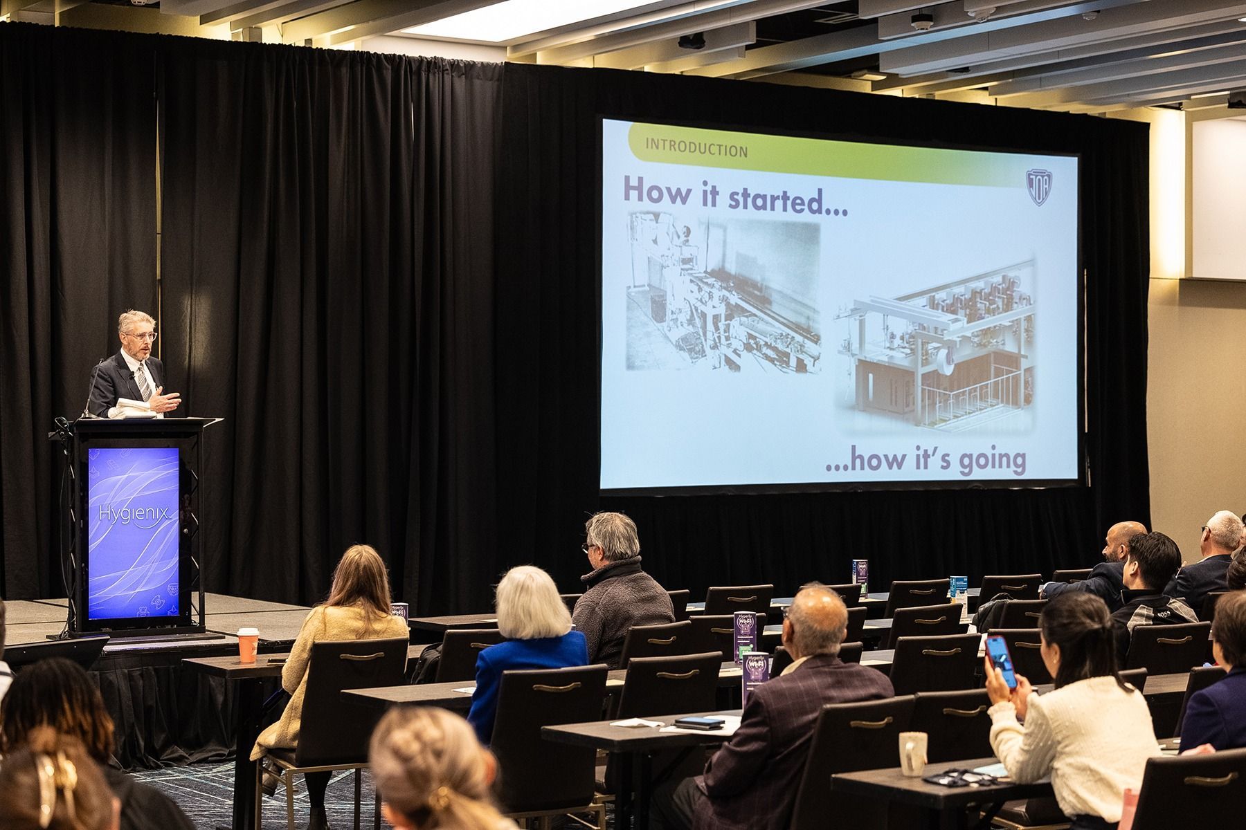 A man lecturing at a conference, screen showing 