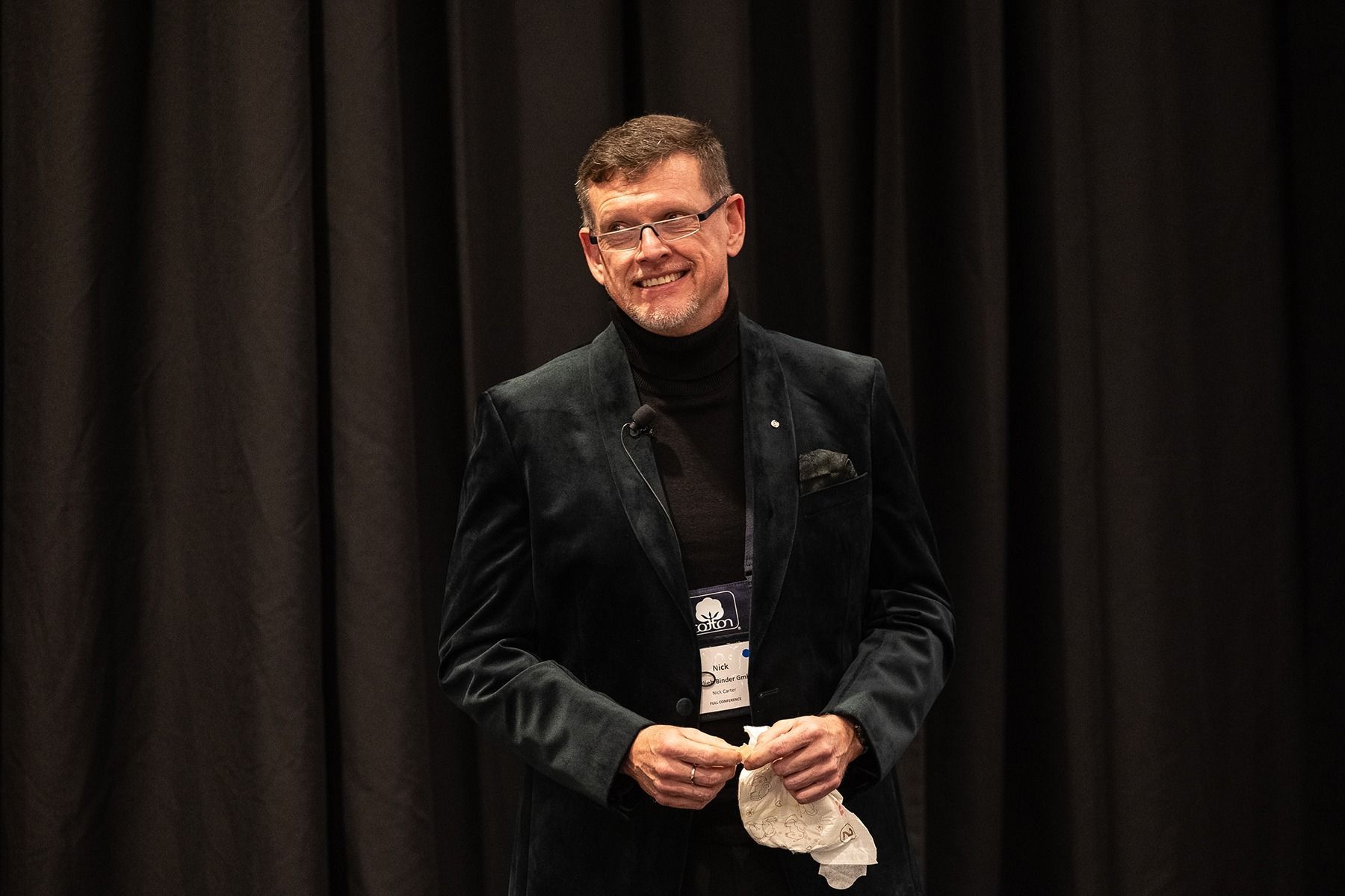 Man in black turtleneck and velvet jacket smiles, standing in front of a dark curtain, holding a white cloth.