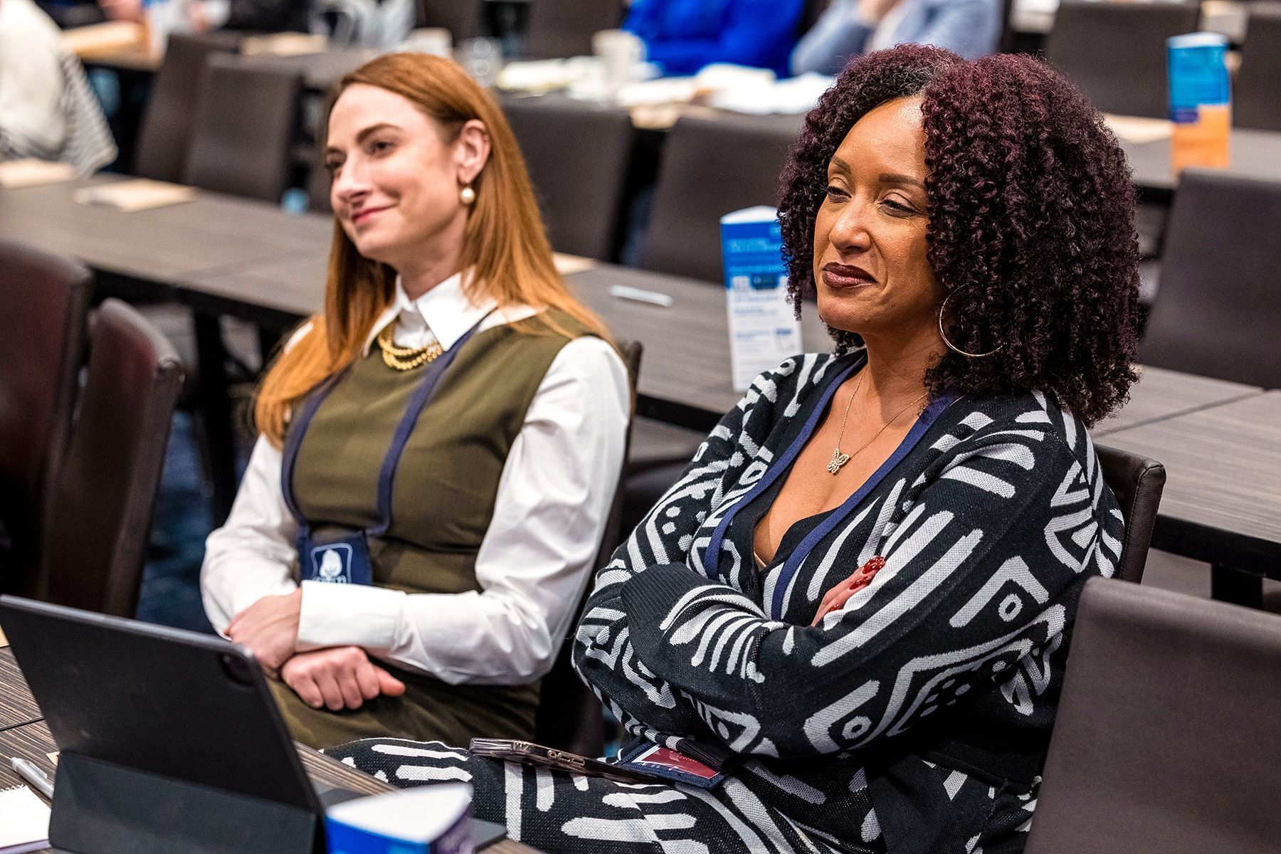 Two women seated at a conference table, listening. One wears a patterned sweater, the other a vest and blouse.
