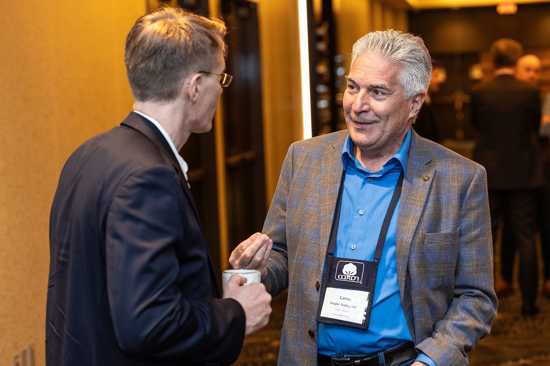 Two men in a hallway, one smiling, holding a cup. Man in a blue shirt and patterned blazer gestures.