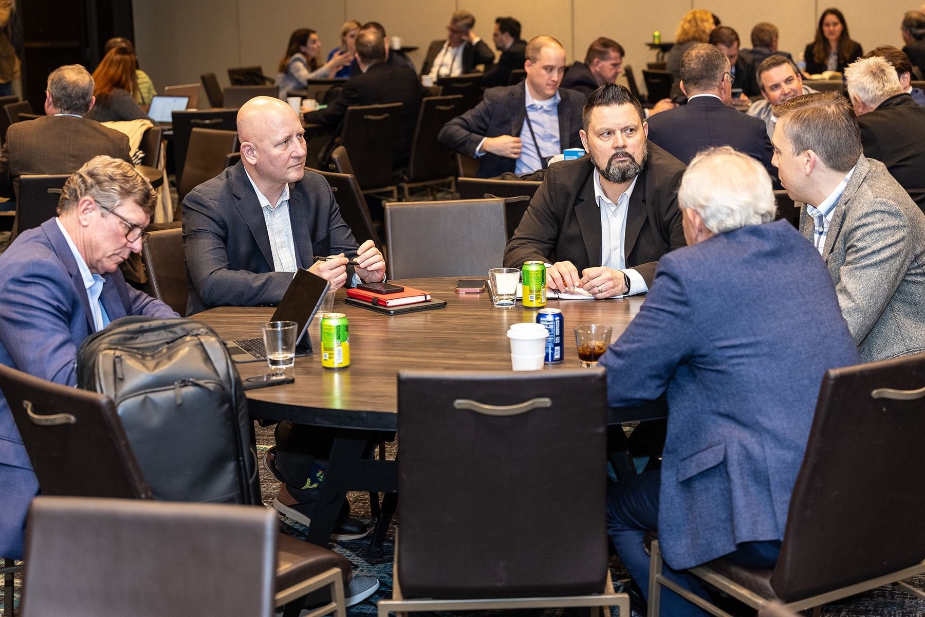 Group of people in suits seated around a round table, conversing. Some have drinks and a laptop.