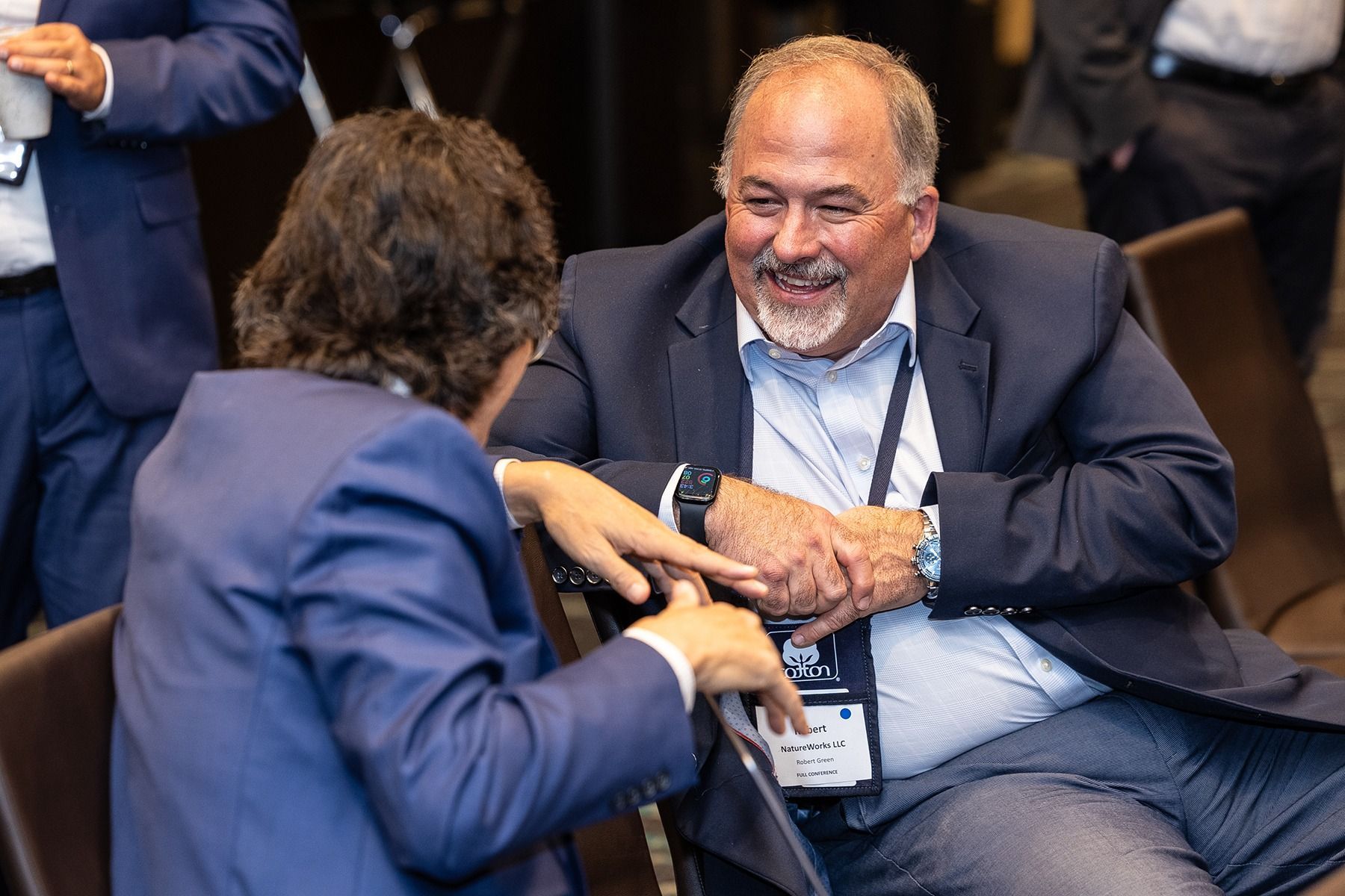 Two men in suits seated and conversing, one smiling and gesturing.