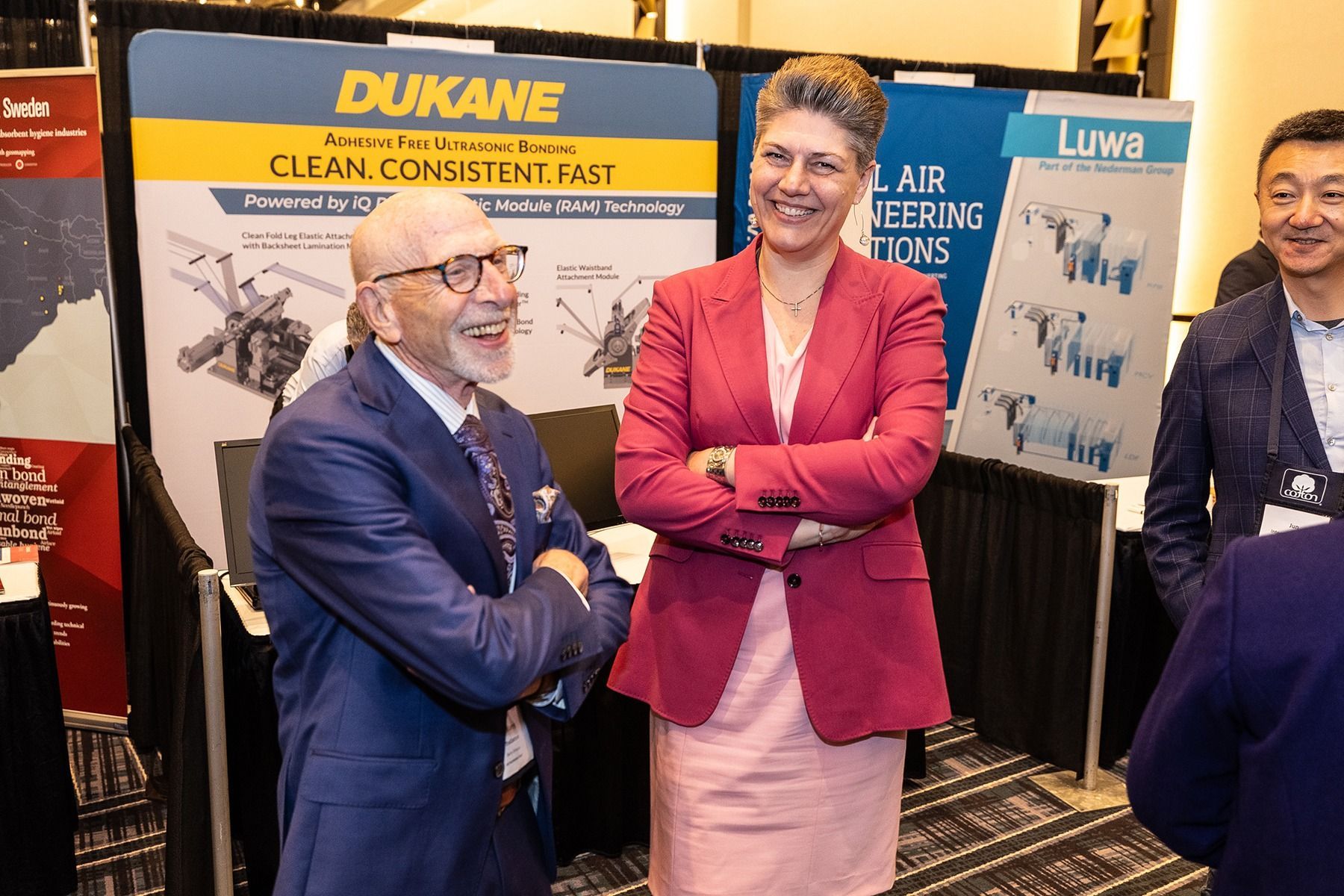 Two people at a trade show booth, smiling. Woman in pink blazer, man in suit with beard. 