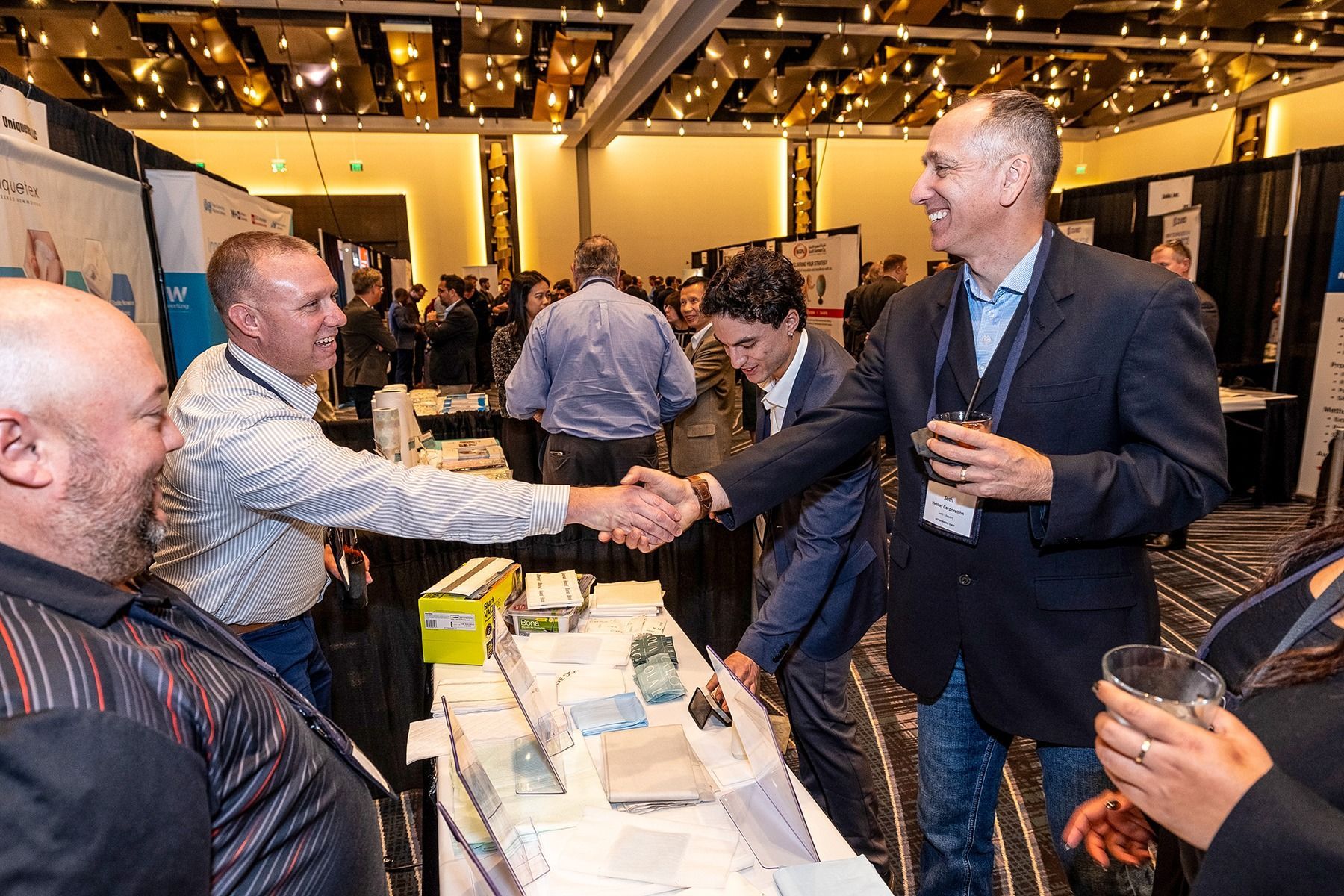 Men in suits shaking hands at a conference booth, smiling. Attendees in background.