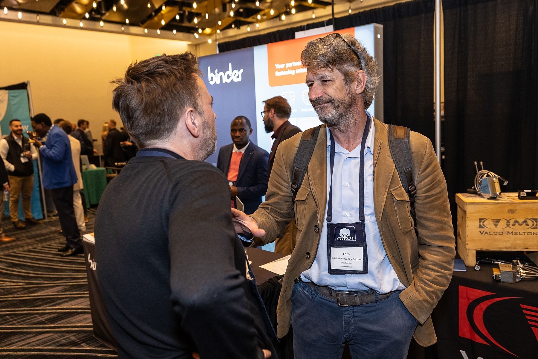 Two men talking at a conference booth; one in a brown jacket, name tag. Others in the background.