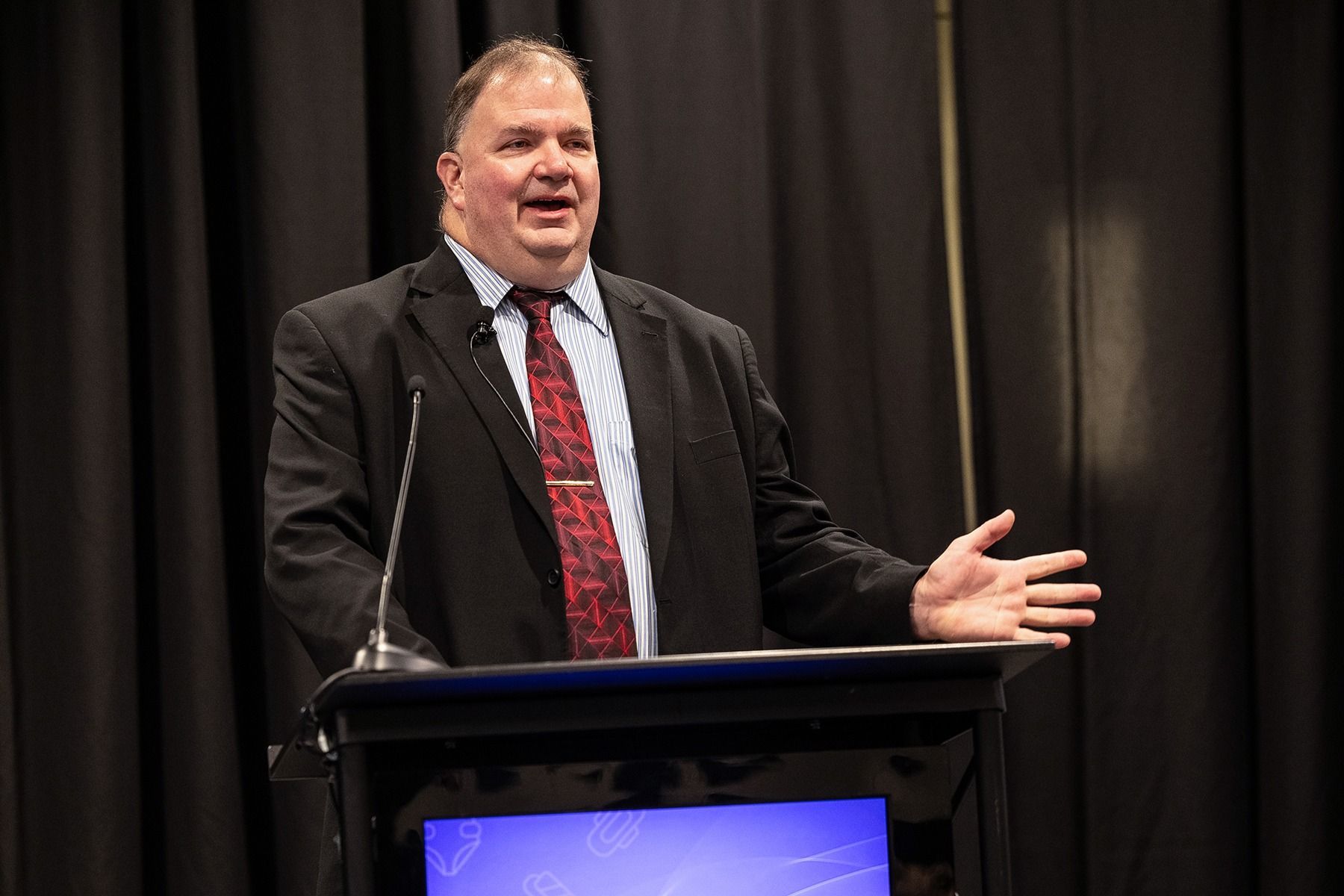 Man in suit speaking at a podium, gesturing with his hand. Dark background.