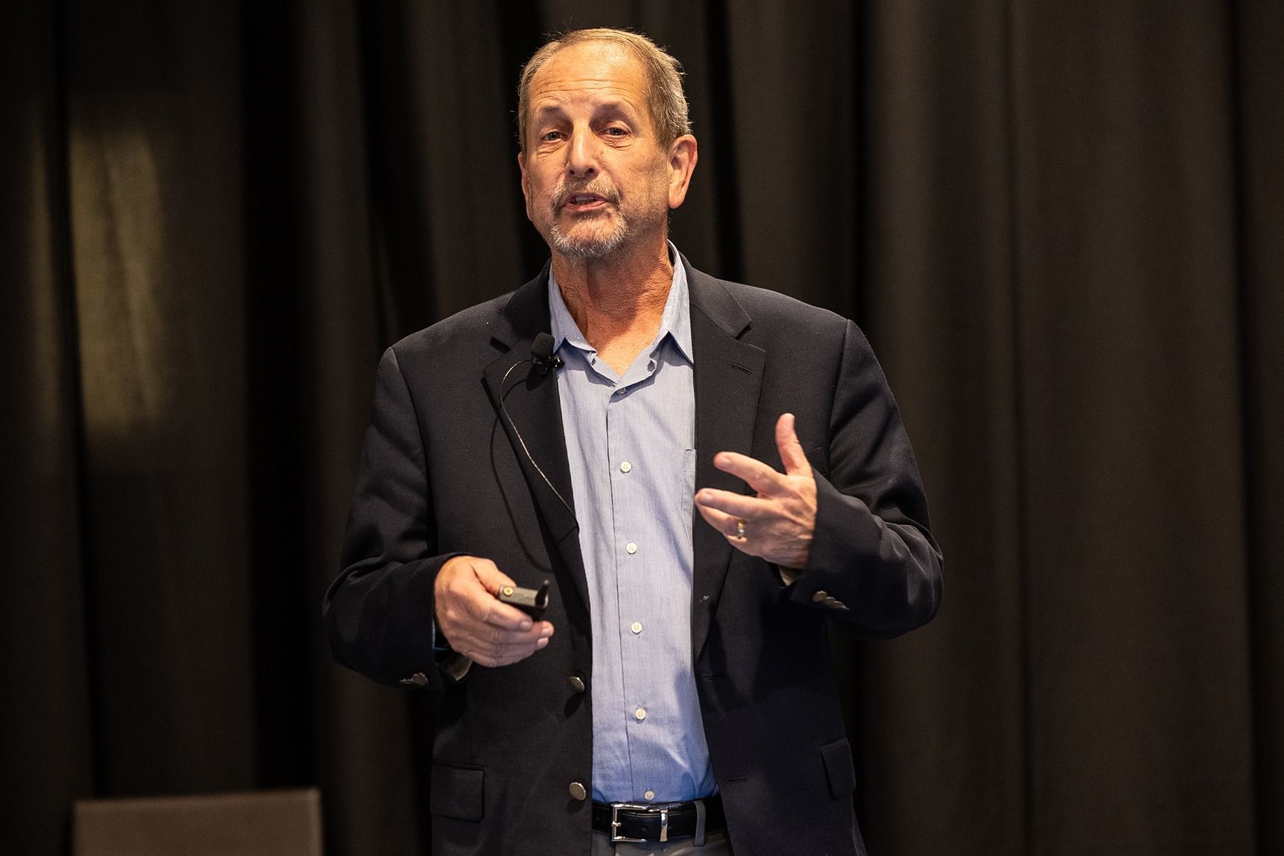 Man in blazer and open shirt speaking at a conference, gesturing with his hands. Dark background.