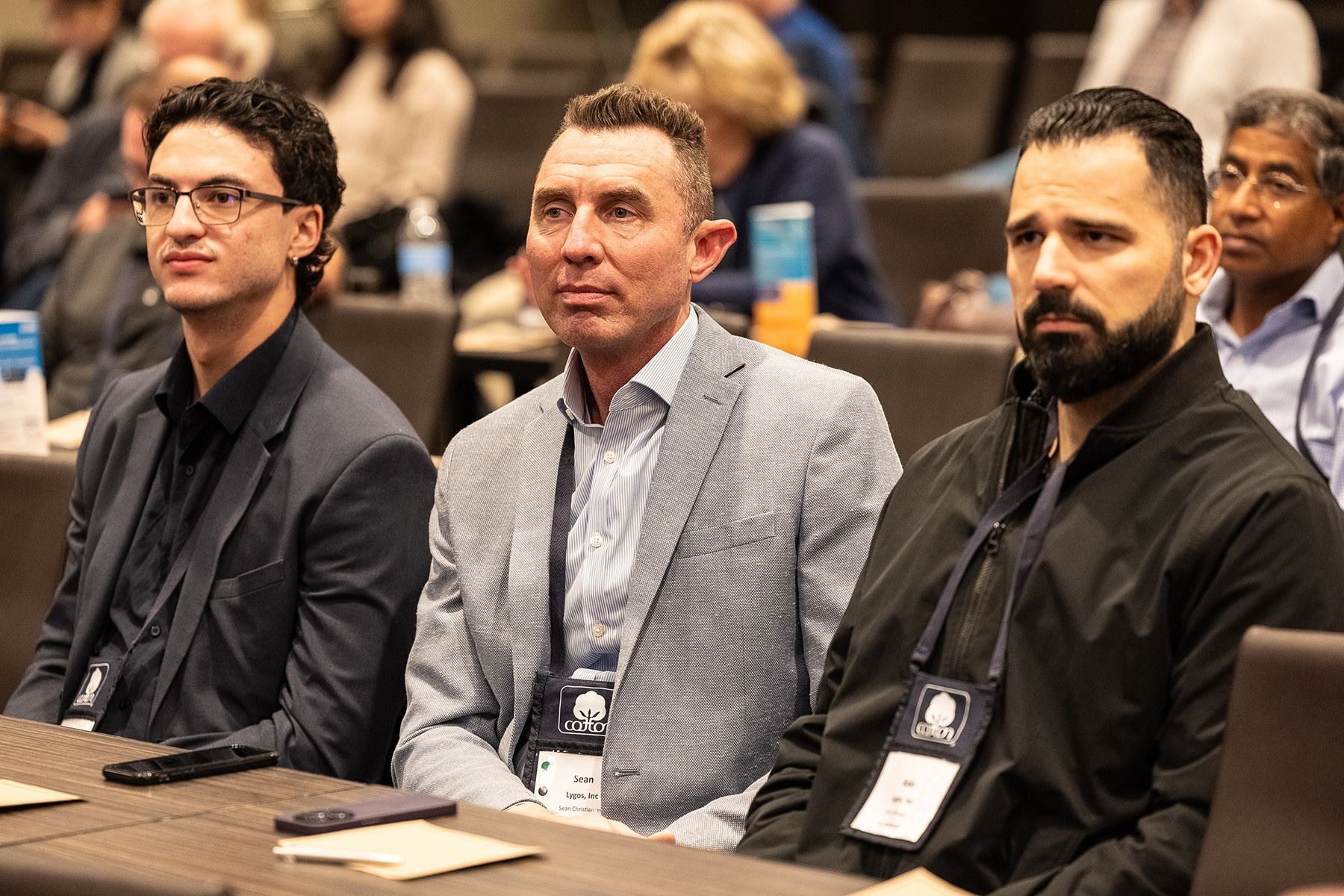 Three men sit at a conference table, listening attentively. Two are wearing jackets, one with glasses.