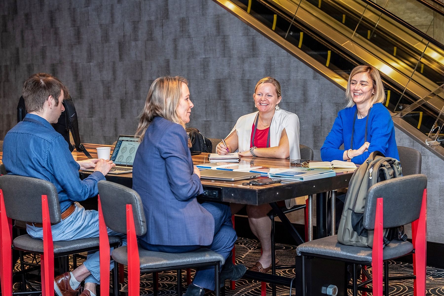 Four people sit around a table in a room with a staircase. They are discussing and looking at items on the table.
