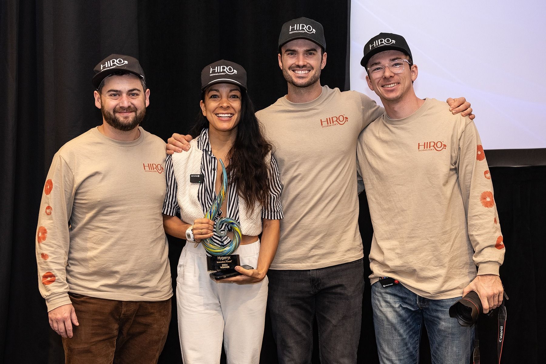 Four people pose, smiling, holding an award on a stage. They wear matching hats and shirts; the woman in the center holds the award.