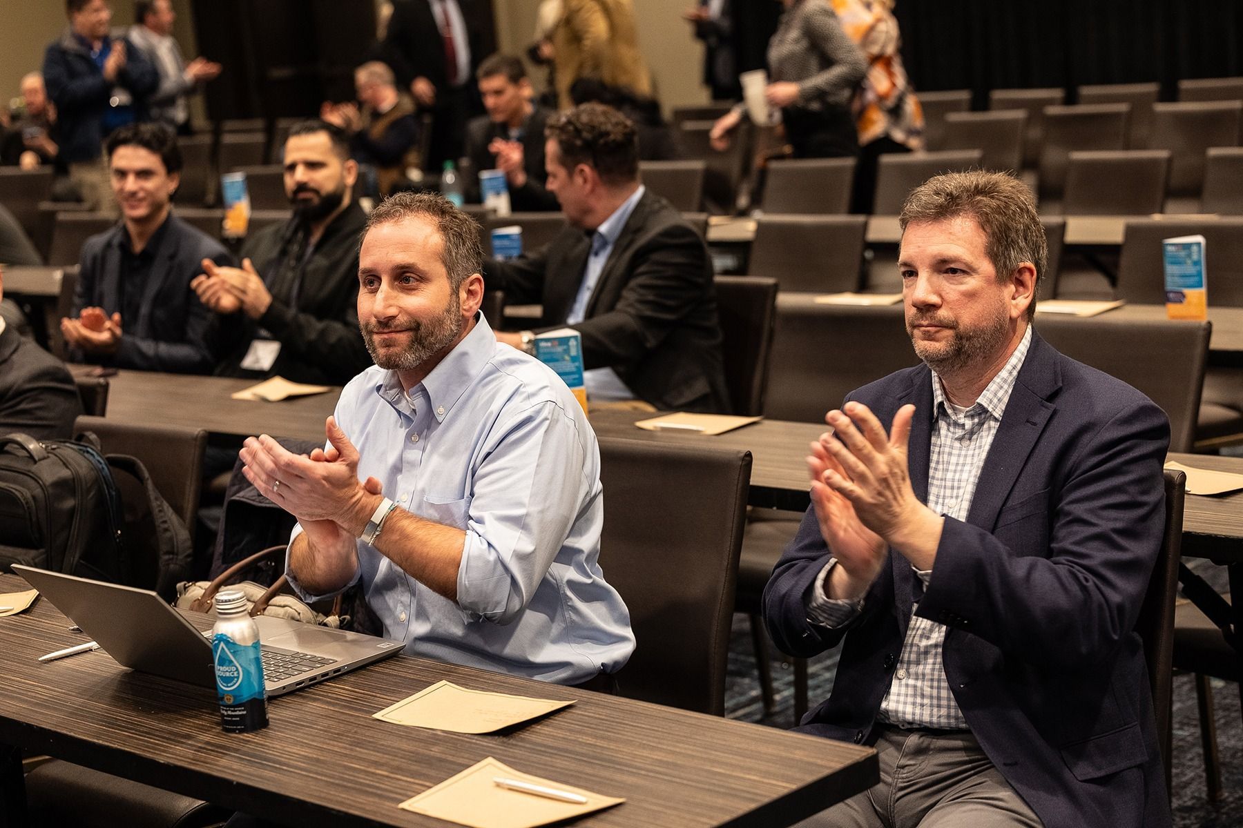 Attendees clapping at a conference, sitting at tables with laptops, in a well-lit room.