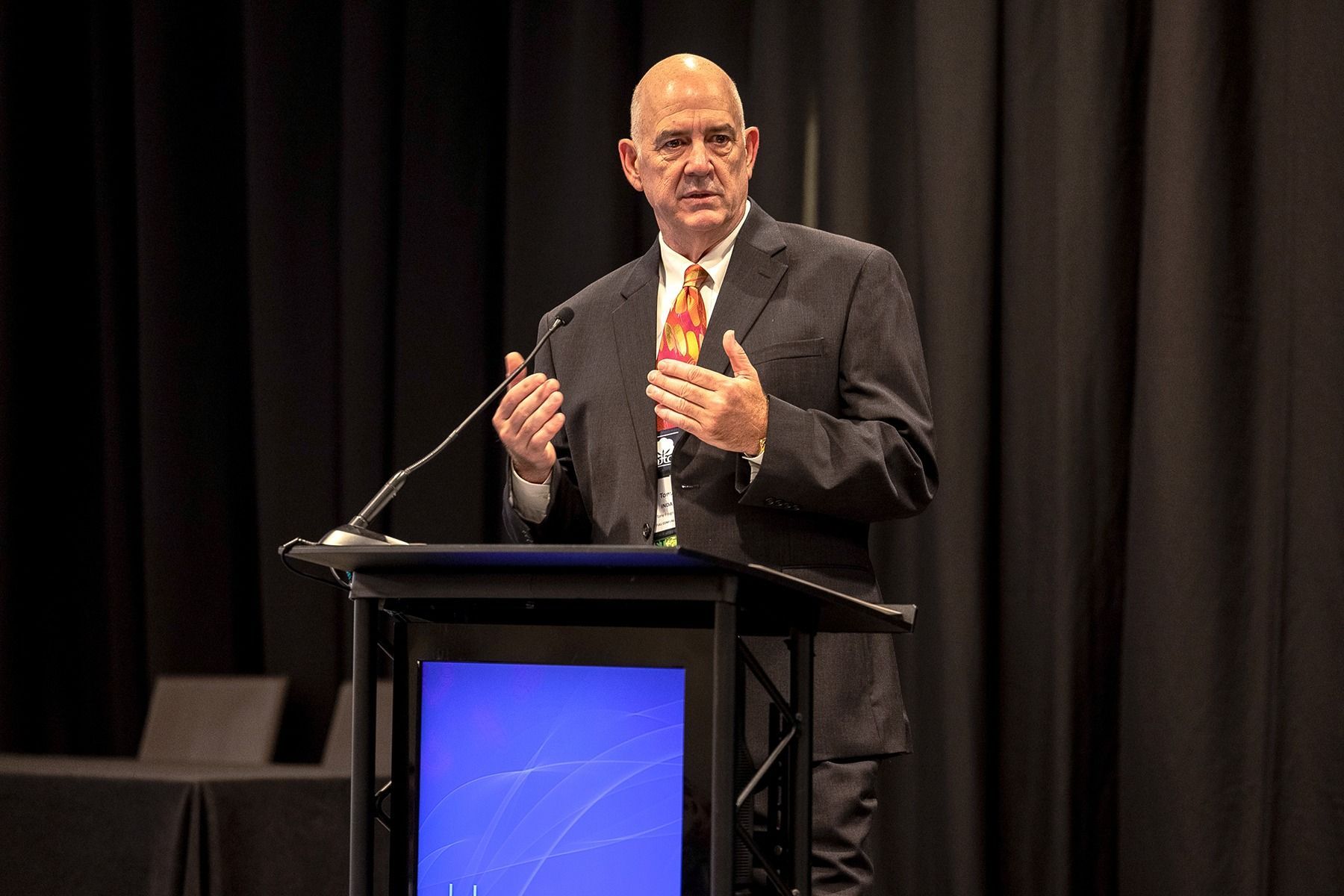 Man in suit speaking at a podium with black curtains in the background.