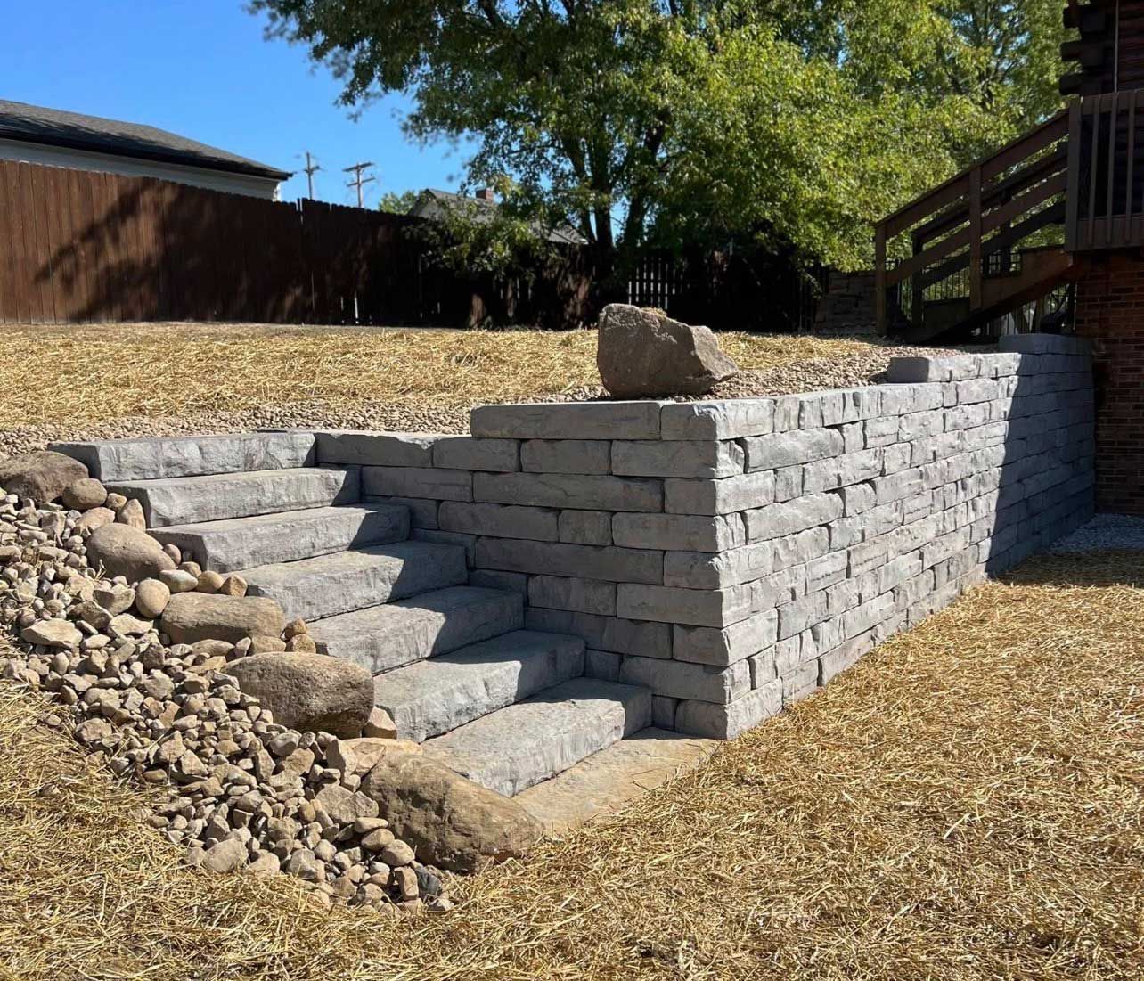 A stone retaining wall with integrated steps leading up a grassy slope near a wooden deck.