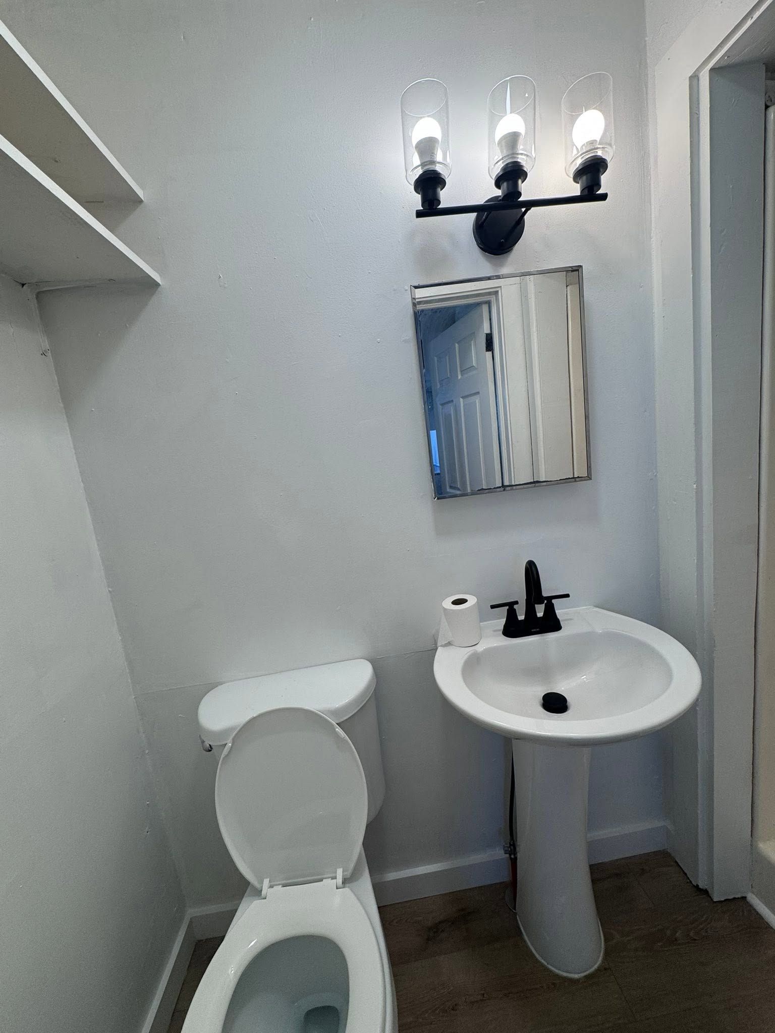 Small bathroom with toilet, pedestal sink, mirror, and three-light wall fixture.