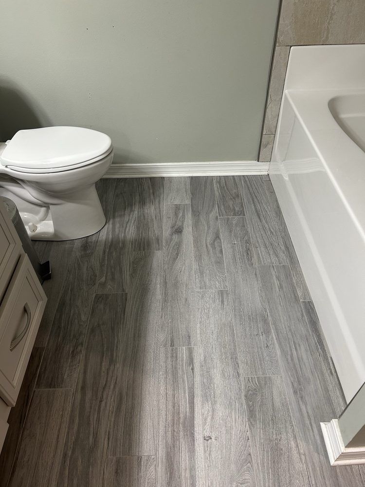 Small bathroom with toilet, gray wood-look floor, white tub, and gray wall.