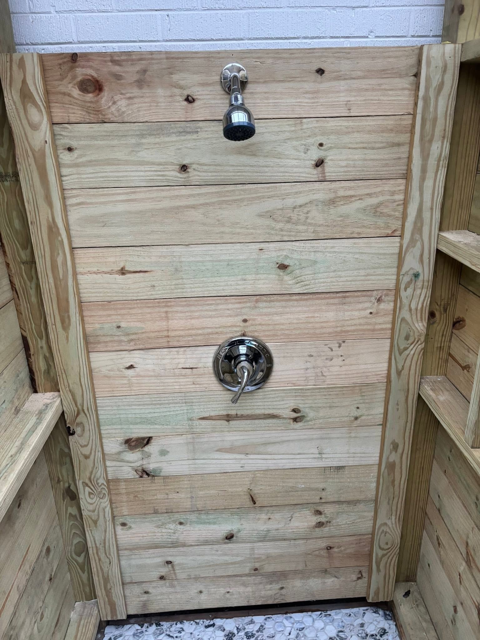 Wooden outdoor shower wall with a faucet and round knob, flanked by railings and a gray brick wall.