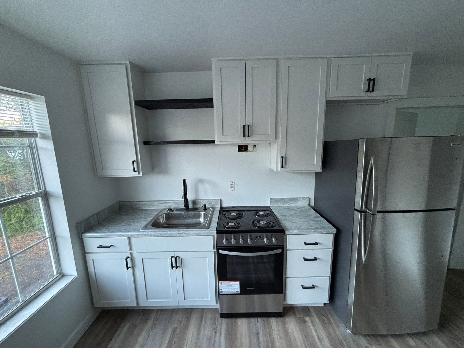 Small white kitchen with cabinets, stove, sink, and stainless steel fridge next to a window.