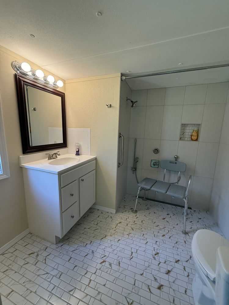 Small bathroom with white tile, vanity and mirror lights, and a walk-in shower area with a chair.