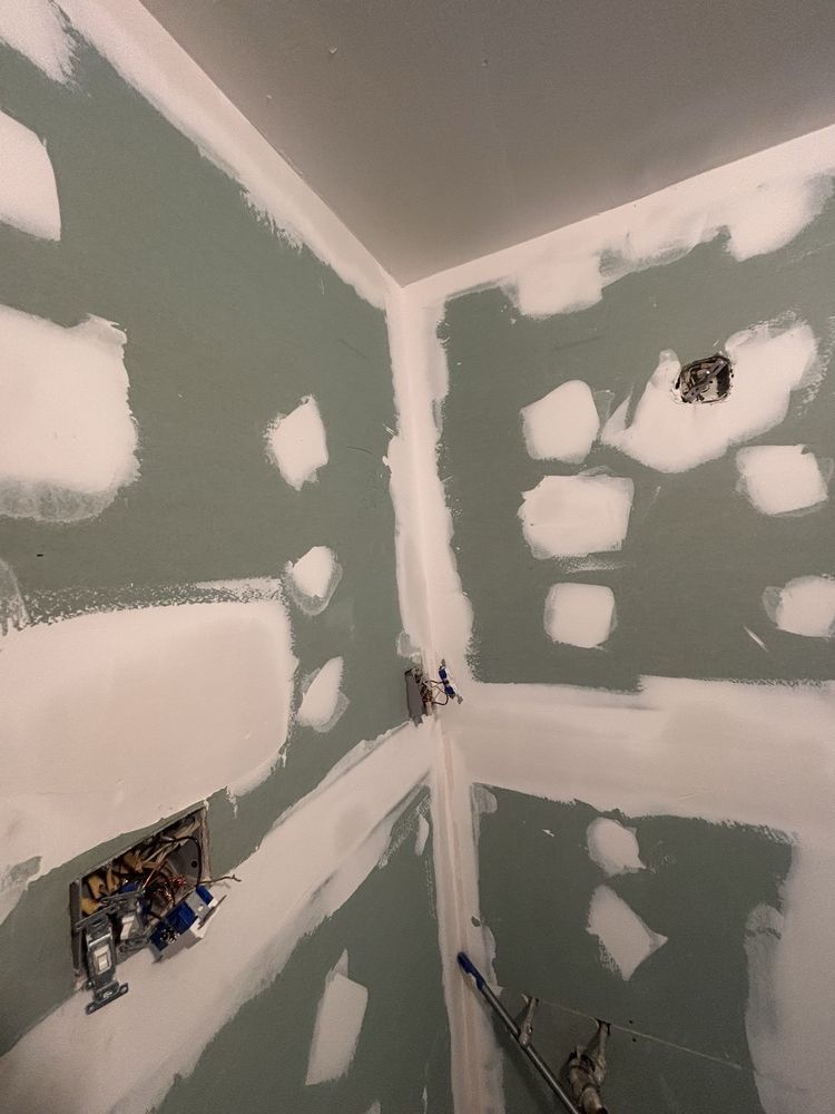 Unfinished bathroom corners with green drywall, white joint compound patches, and exposed electrical boxes.