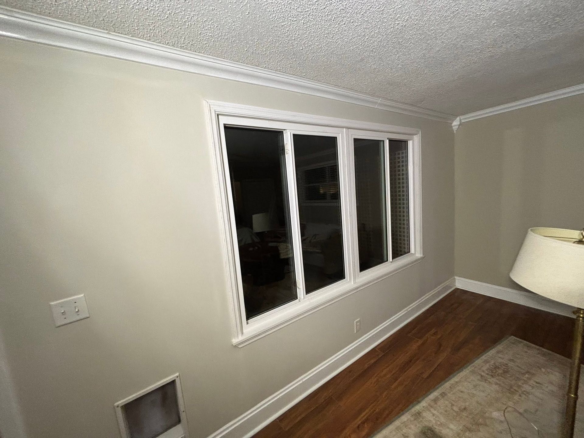 Empty living room with beige walls, hardwood floor, large window, and a rug on the right