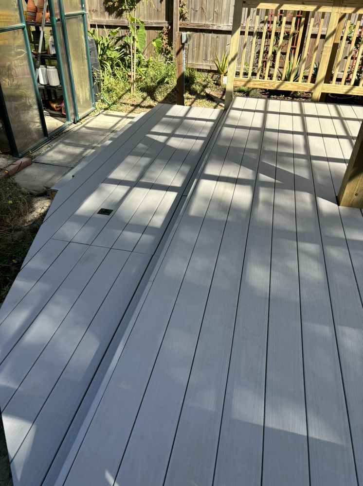 Light gray wooden deck with long shadows, bordered by a railing and garden plants.