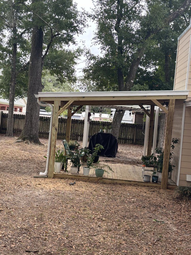 Backyard patio with a pergola, potted plants, a grill, and large trees behind a wooden fence.