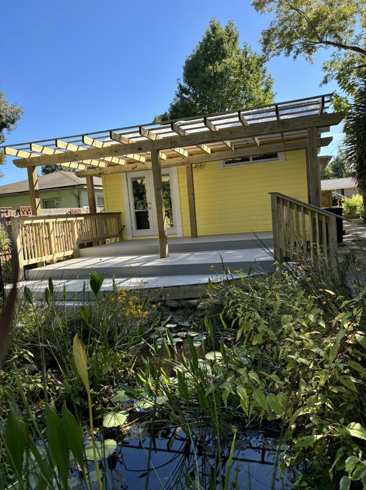 Yellow cottage with wooden porch and pergola beside a pond, surrounded by plants under a blue sky