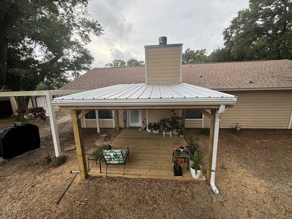 Back porch with white roof, beige house, potted plants, and gravel yard under cloudy sky