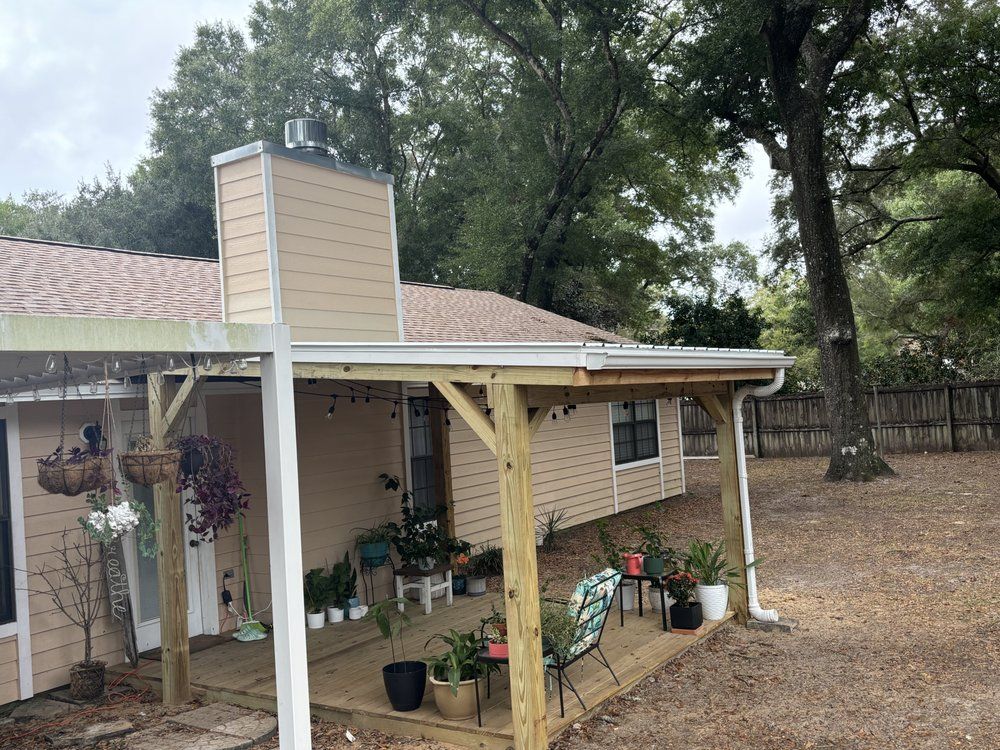 Small beige house porch with white posts, hanging plants, and potted flowers under tall trees